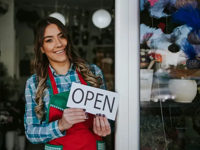 Smiling small business owner at shop doorway holding an OPEN sign, inviting customers and promoting local listings